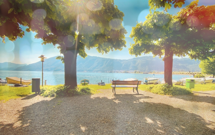 Sonniger Uferbereich im Süden der Schweiz mit Blick auf einen See, schattenspendenden Bäumen, Sitzbank und Bergen im Hintergrund – warme Lichtstimmung mit Bokeh-Effekt.