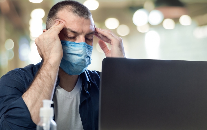 Erschöpfter Mann mit Schutzmaske sitzt gestresst vor dem Laptop und symbolisiert psychische Belastung sowie Präsentismus im Arbeitsalltag